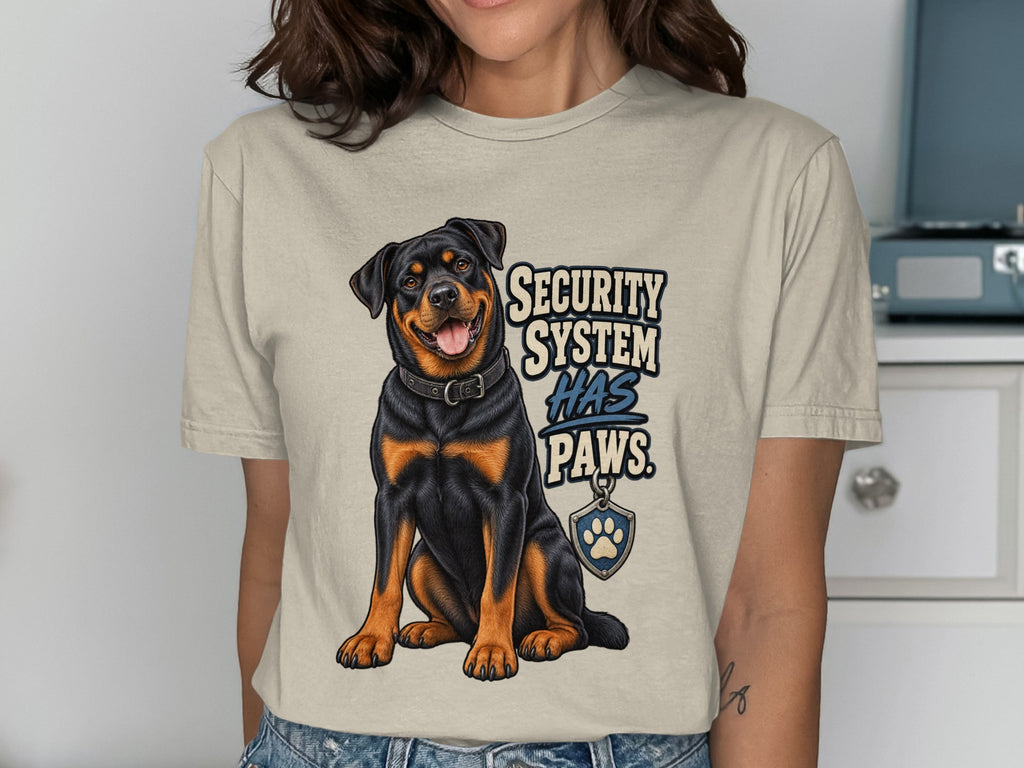 Rottweiler dog illustration with text "SECURITY SYSTEM HAS PAWS" on a cream t-shirt.