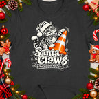 Dark t-shirt with a tabby cat wearing a Santa hat, holding an orange cone, and text "Santa Claws".